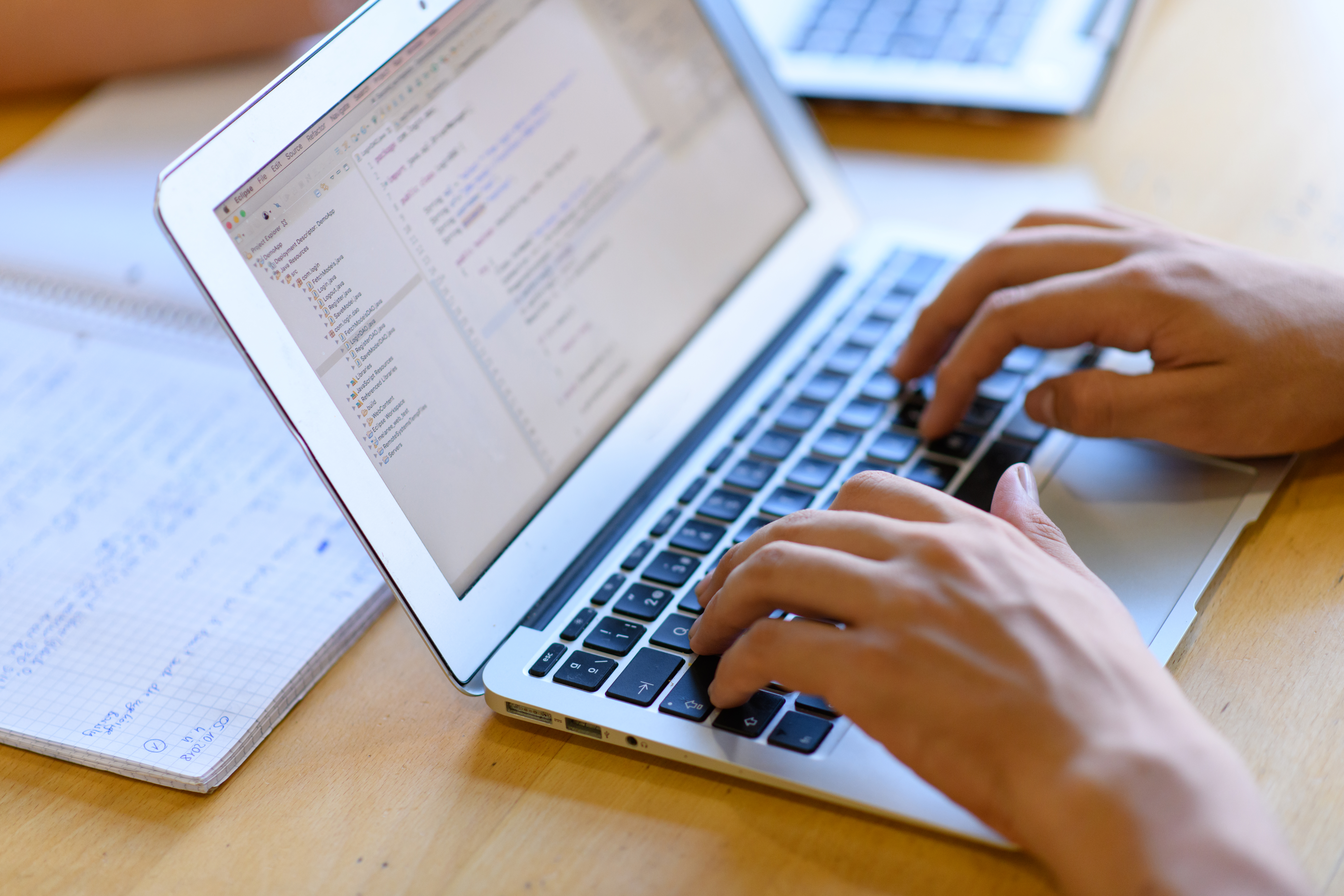Eine Person, deren Hände im Bild sind, schreibt an einem Laptop. Hinter dem Laptop liegt ein aufgeschlagener Collegeblock mit handschriftlichen Einträgen.