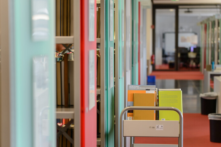 Ein Bücherwagen mit Büchern steht vor Bücherregalen mit grünen und roten Regalfronten im Bibliotheksbereich Schloss Ehrenhof.