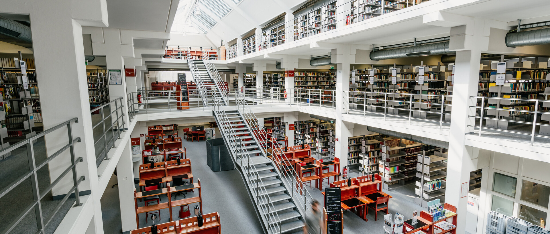 Bibliotheksbereich A3 mit vielen Arbeitsplätzen und Bücherregalreihen im Erdgeschoss, dem ersten und zweiten Stock mit Bücherregalreihen, einer langen Treppe vom Erdgeschoss bis in den zweiten Stock und einem sonnigen Glasdach.