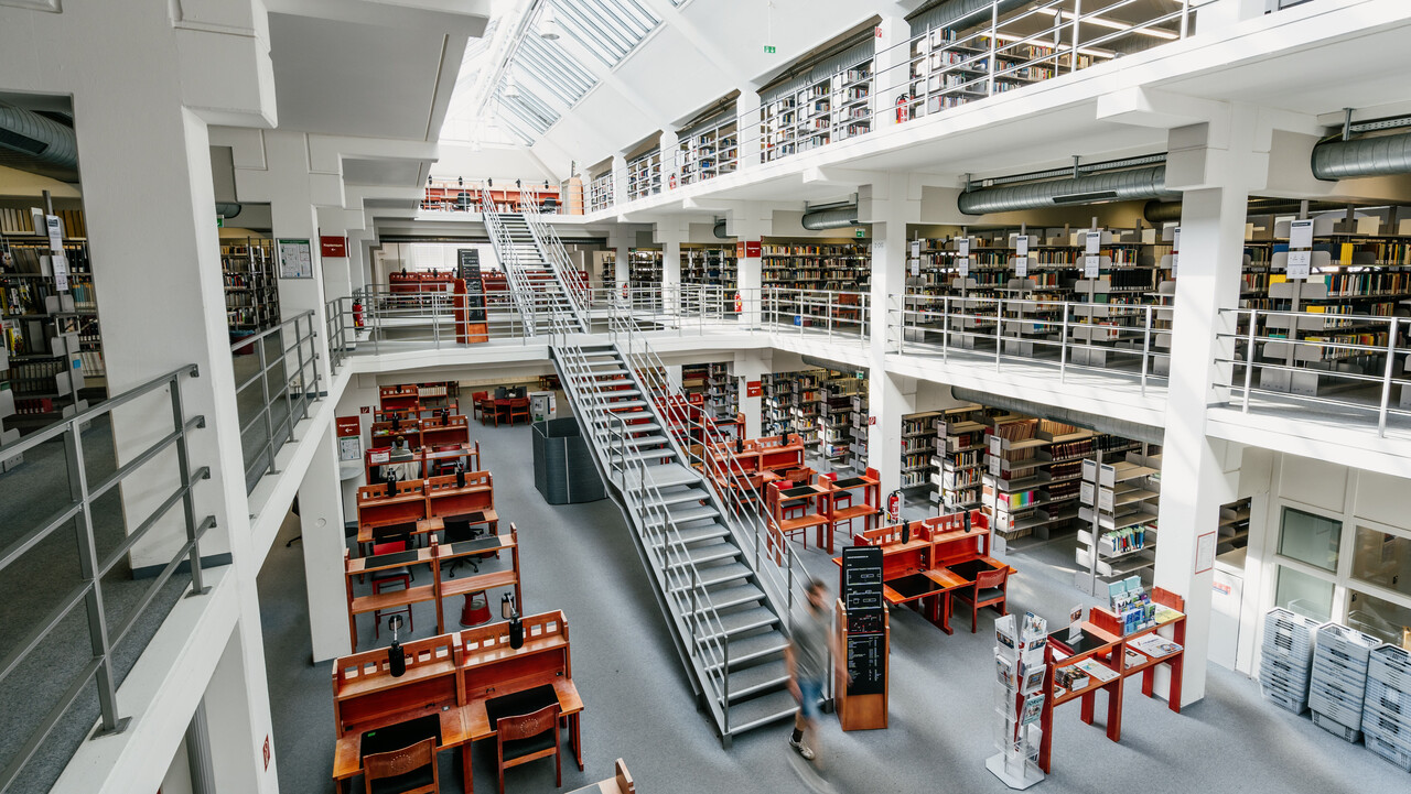 Bibliotheksbereich A3 mit vielen Arbeitsplätzen und Bücherregalreihen im Erdgeschoss, dem ersten und zweiten Stock mit Bücherregalreihen, einer langen Treppe vom Erdgeschoss bis in den zweiten Stock und einem sonnigen Glasdach.