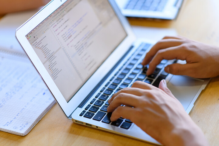 Eine Person, deren Hände im Bild sind, schreibt an einem Laptop. Hinter dem Laptop liegt ein aufgeschlagener Collegeblock mit handschriftlichen Einträgen.