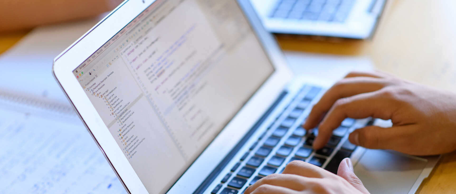 Eine Person, deren Hände im Bild sind, schreibt an einem Laptop. Hinter dem Laptop liegt ein aufgeschlagener Collegeblock mit handschriftlichen Einträgen.