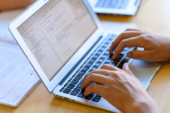 Eine Person, deren Hände im Bild sind, schreibt an einem Laptop. Hinter dem Laptop liegt ein aufgeschlagener Collegeblock mit handschriftlichen Einträgen.