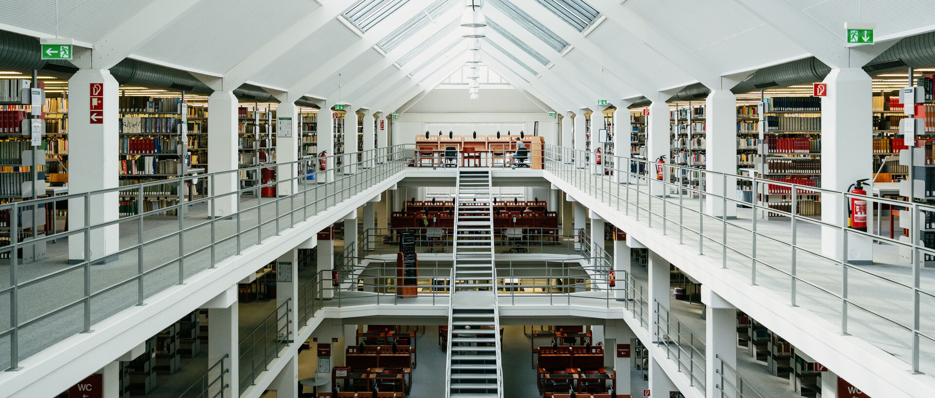 Bibliotheksbereich A3 mit Arbeitsplätzen im Erdgeschoss, dem ersten und zweiten Stock mit Bücherregalreihen und langer Treppe vom Erdgeschoss in den zweiten Stock mit sonnigem Glasdach.
