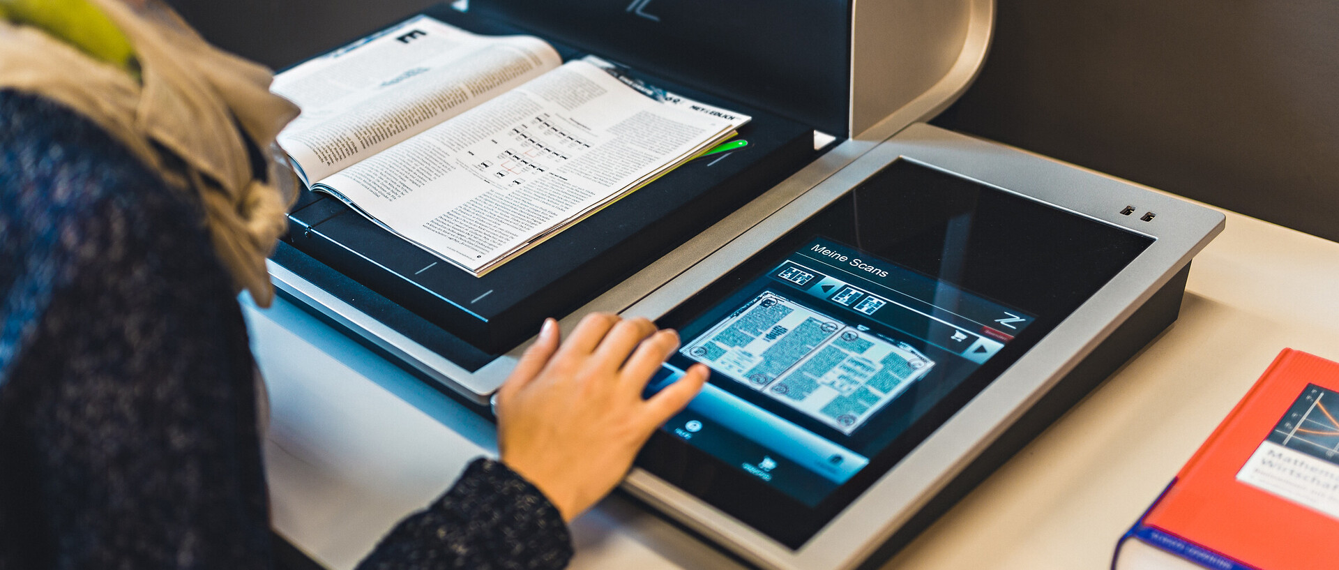 Eine Studentin benutzt einen Scanner in der Bibliothek. Ein Buch liegt auf der Scanfläche und sie nimmt die Einstellungen am Bedienfeld vor. Ein weiteres Buch liegt rechts neben dem Scanner auf dem Tisch.