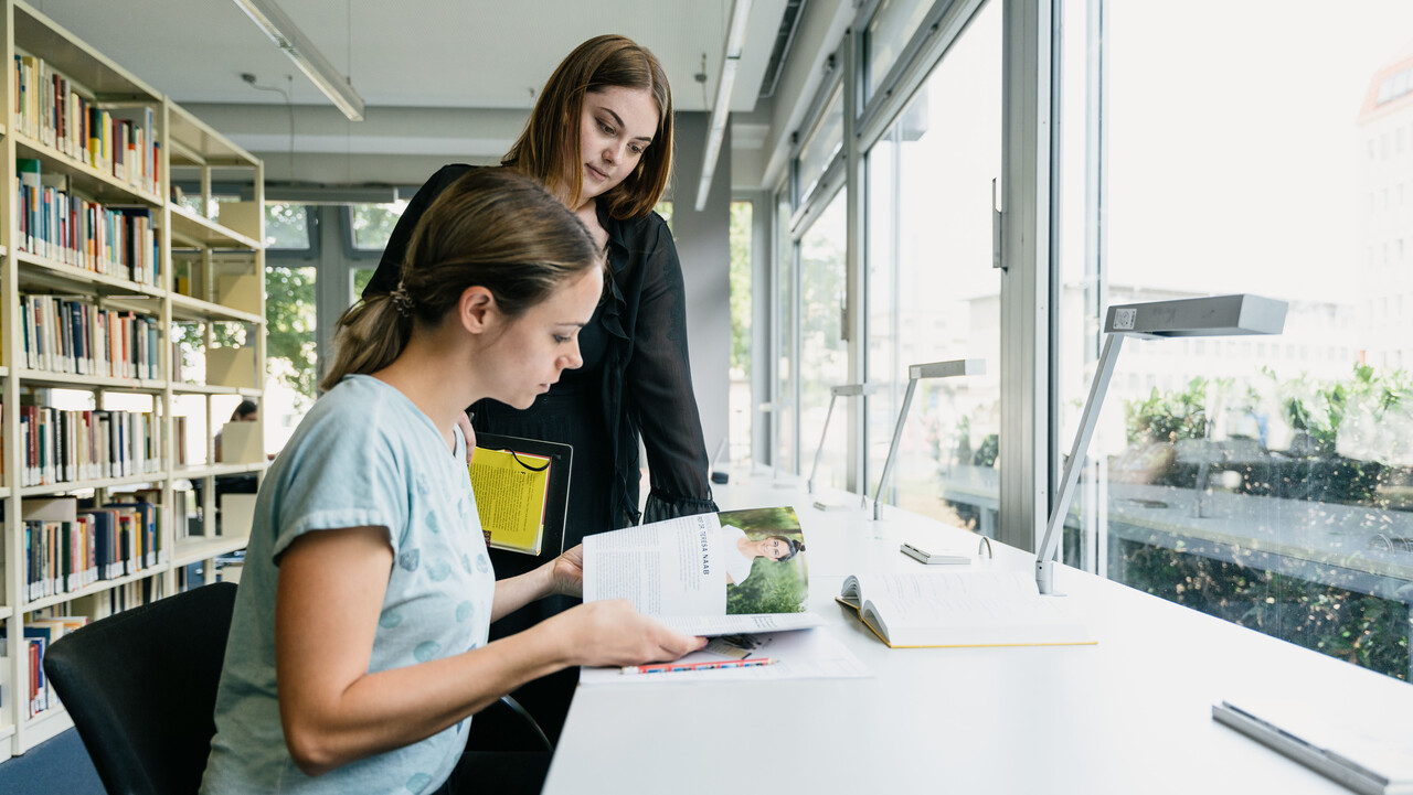 Zwei junge Frauen sehen sich gemeinsam an einem Tisch am Fenster im Bibliotheksbereich A5 eine Zeitschrift an.