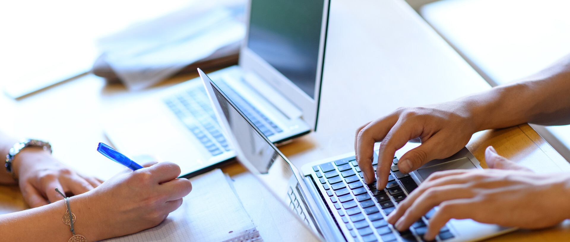 Zwei Personen sitzen sich an einem Tisch gegenüber, es sind jeweils nur die Hände zu sehen. Die links sitzende Person schreibt in einen Collegeblock, dahinter steht ein aufgeklapptes Laptop. Die rechts sitzende Person schreibt an einem weiteren Laptop.