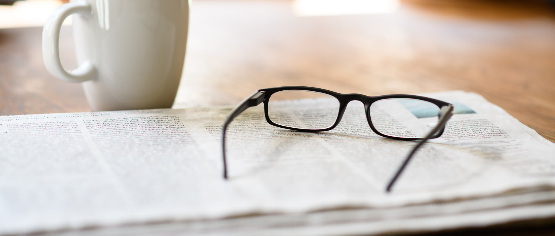 Eine Zeitung auf der eine Lesebrille liegt. Daneben steht eine Kaffeetasse