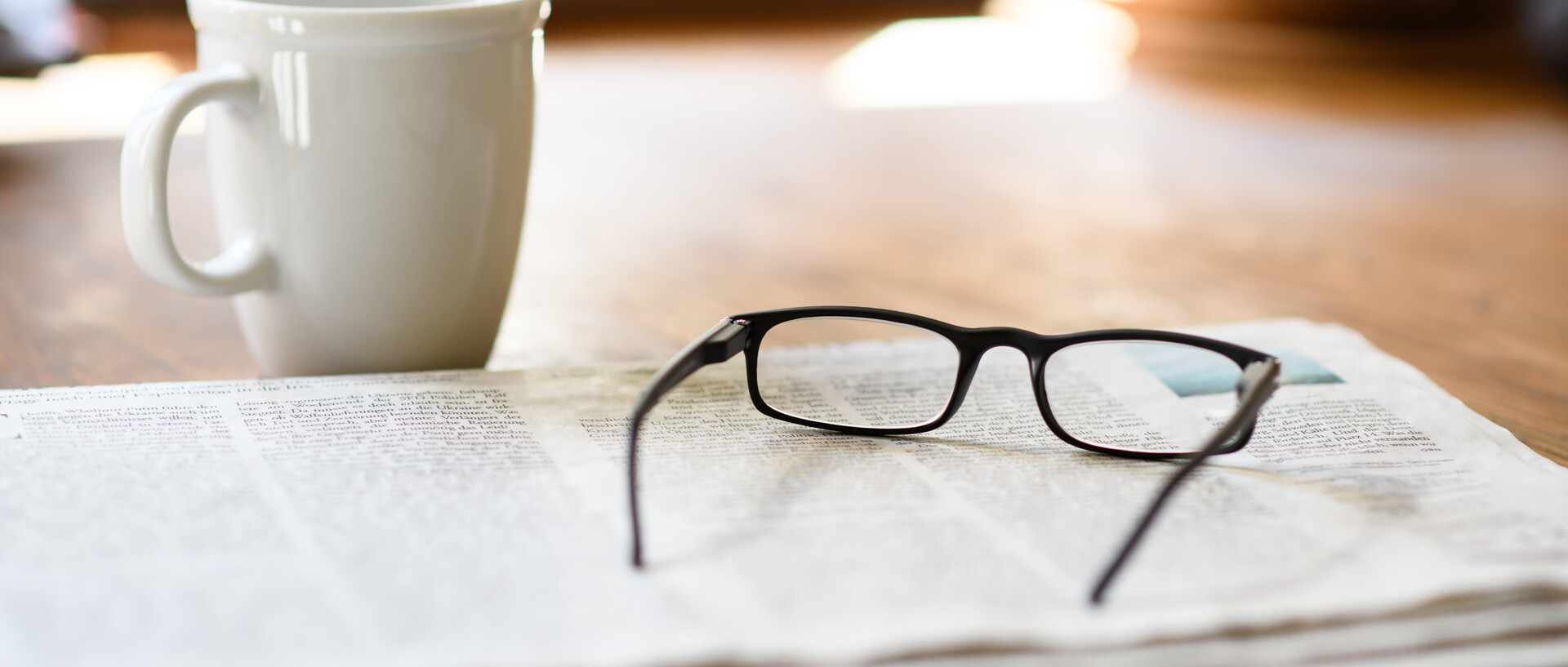 A newspaper with a pair of reading glasses on top. Next to it is a coffee cup.