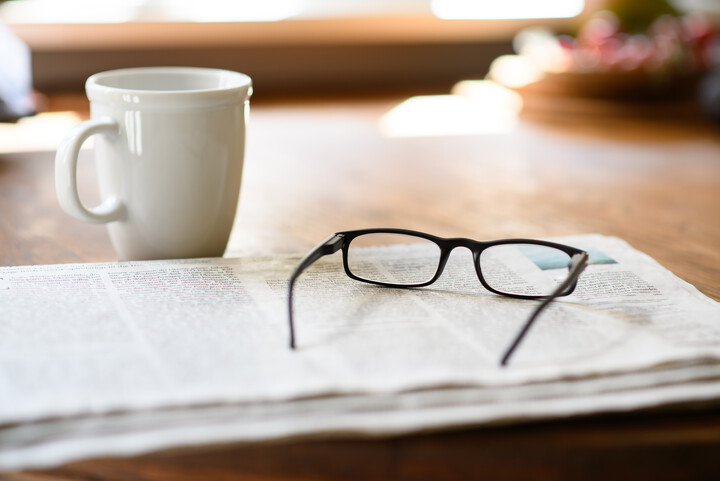 Eine Zeitung auf der eine Lesebrille liegt. Daneben steht eine Kaffeetasse